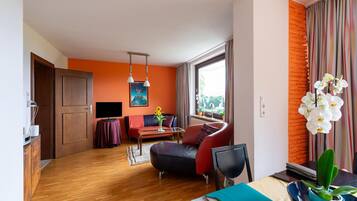 A modern hotel room with a wooden floor, a sofa, a coffee table, a TV, and a window with curtains.