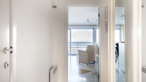 A hotel corridor with a view of the sea through large windows, a white door with a keyhole, and a radiator on the wall. - Les Petrels 302 on the Beach in Koksijde (Koksijde)