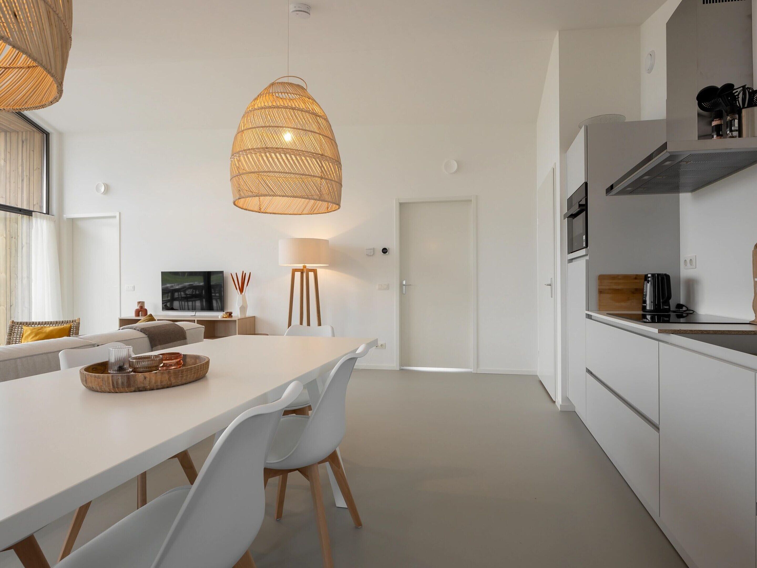 A modern kitchen with white cabinetry, a dining area with a long table and white chairs, and a large woven pendant light.