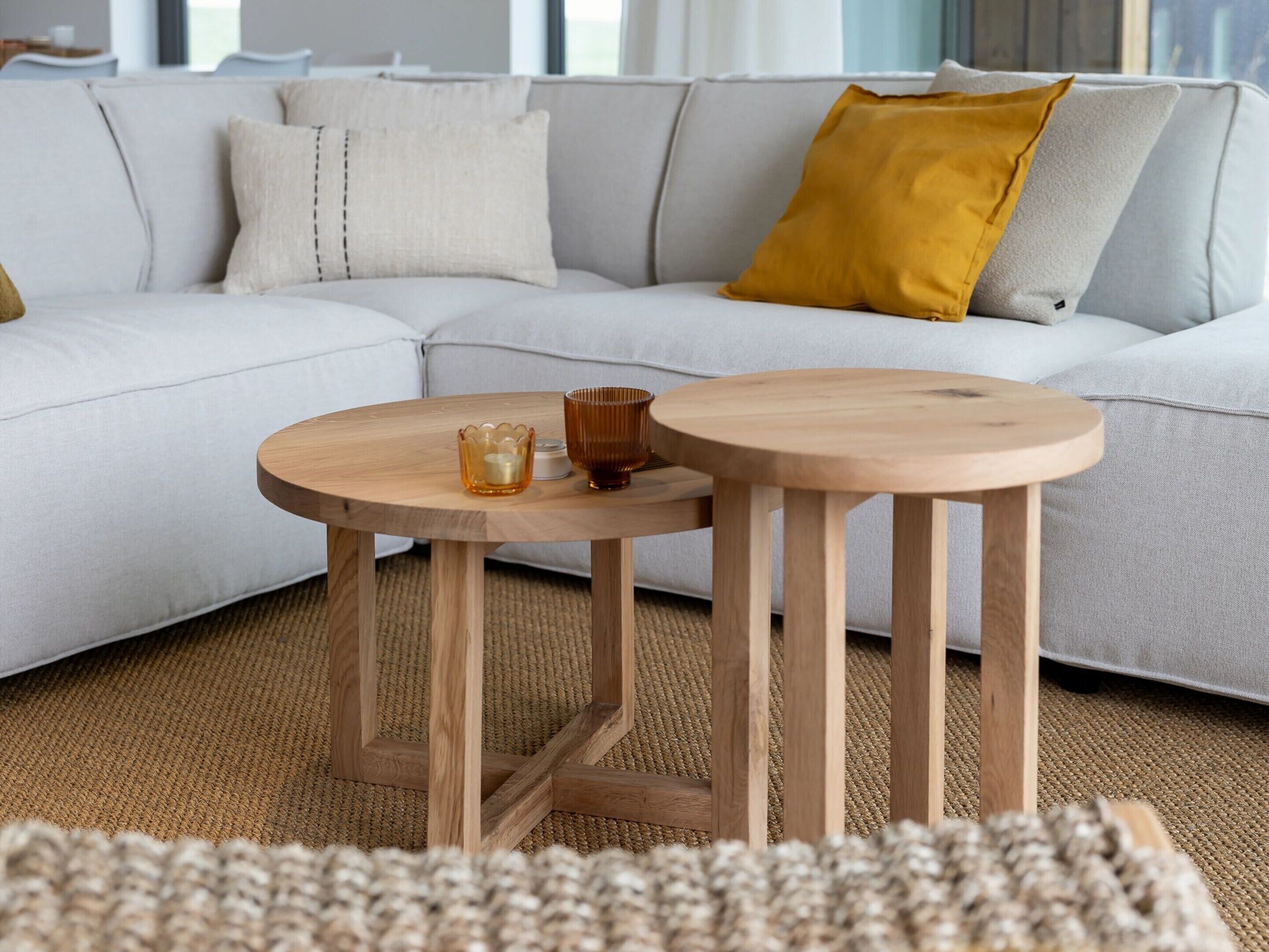 Ein modernes Wohnzimmer mit einem hellgrauen Sofa, einem senfgelben Kissen und zwei Holztischen mit Kerzen.