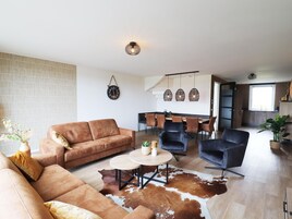 Una sala de estar moderna con un sofá de cuero marrón, una mesa de centro redonda y un tapiz de piel de vaca. Hay un comedor con una mesa y sillas de madera, y una cocina con armarios de madera y una ventana.