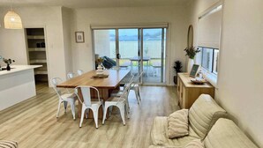 Ein modernes Wohnzimmer mit einem Holzessttisch, weißen Stühlen, einem beigen Sofa und einer großen Glaswand, die auf einen Balkon führt.