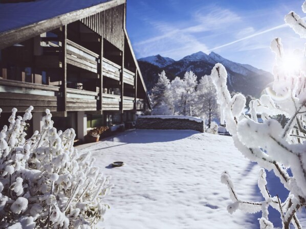 A wooden building with a sloped roof and multiple balconies, surrounded by snow-covered trees and mountains in the background.