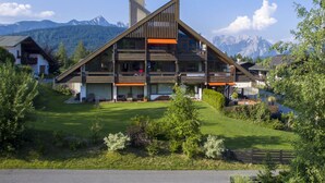 A modern multi-story building with a sloped roof, surrounded by lush greenery and mountains in the background. - Dachgeschoss-atelier (Seefeld in Tirol)