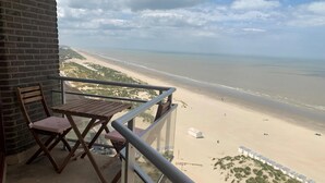 Un balcon avec une chaise et une table en bois, donnant sur une plage au rivage sablonneux et sous un ciel dégagé.