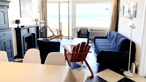 Ein Zimmer mit Schachbrettmuster-Boden, einer Couch, einem Tisch und Stühlen. Ein Fenster bietet Blick auf den Strand.
