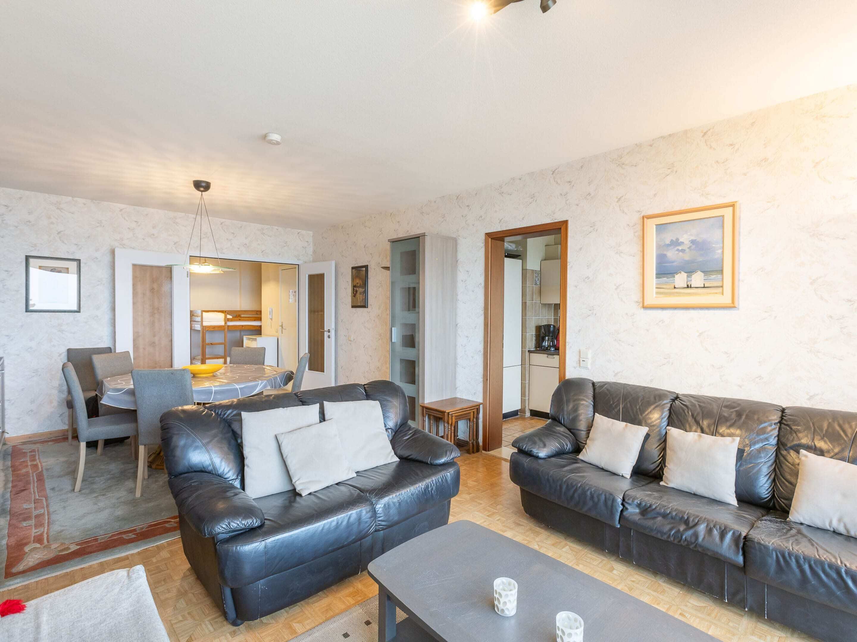 A living room with two black leather sofas, a dining table with chairs, a kitchen area, and a framed picture on the wall.