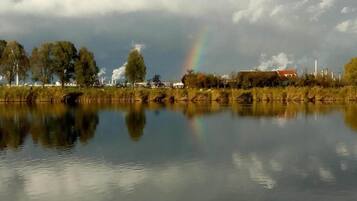 Een rustig meer met een regenboog, bomen en industriële gebouwen op de achtergrond.