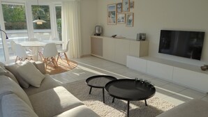A modern living room with a sofa, two coffee tables, a dining area with chairs, a kitchen with a counter, and a TV mounted on the wall. - Apartment in Nieuwpoort With Sea Views (Nieuwpoort)