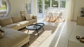 A modern living room with a beige sofa, a coffee table, and a dining area with a white table and chairs. - Apartment in Nieuwpoort With Sea Views (Nieuwpoort)