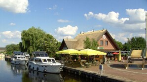 Een steiger aan de rivier met boten, een restaurant met terras en een traditioneel gebouw met rieten dak.