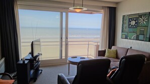 A hotel room with a large sliding glass door leading to a balcony with a view of the beach and ocean. There is a sofa, a chair, and a small table with a remote control on it. - Apartment in Nieuwpoort for 6 Persons (Nieuwpoort)