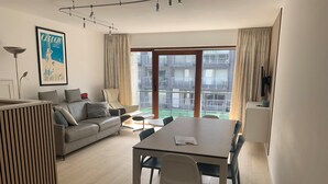 A modern living room with a sofa, a dining table, and a view of the city through large windows. - Apartment in Nieuwpoort Near Marina Views (Nieuwpoort)