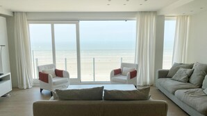 A modern living room with a large sliding glass door leading to a balcony overlooking a beach. There are two armchairs and a sofa with cushions. - Apartment in Nieuwpoort With Ocean Views (Nieuwpoort)