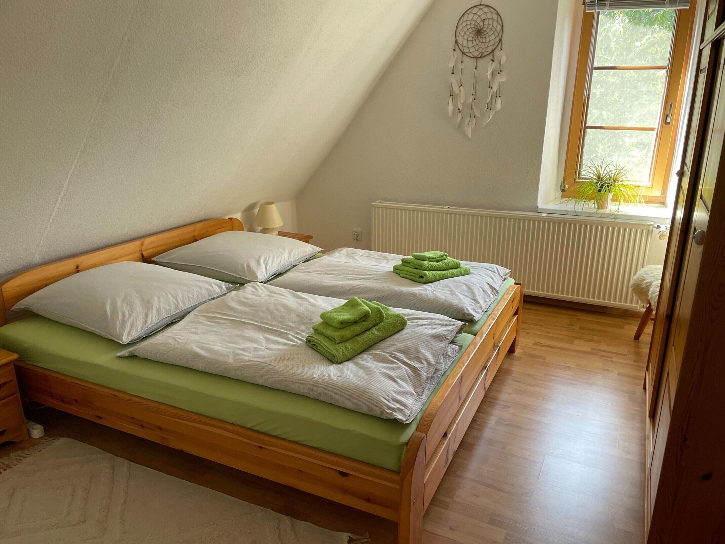 A bedroom with a wooden bed, white pillows, green towels, a dreamcatcher, a window with plants, and a radiator.