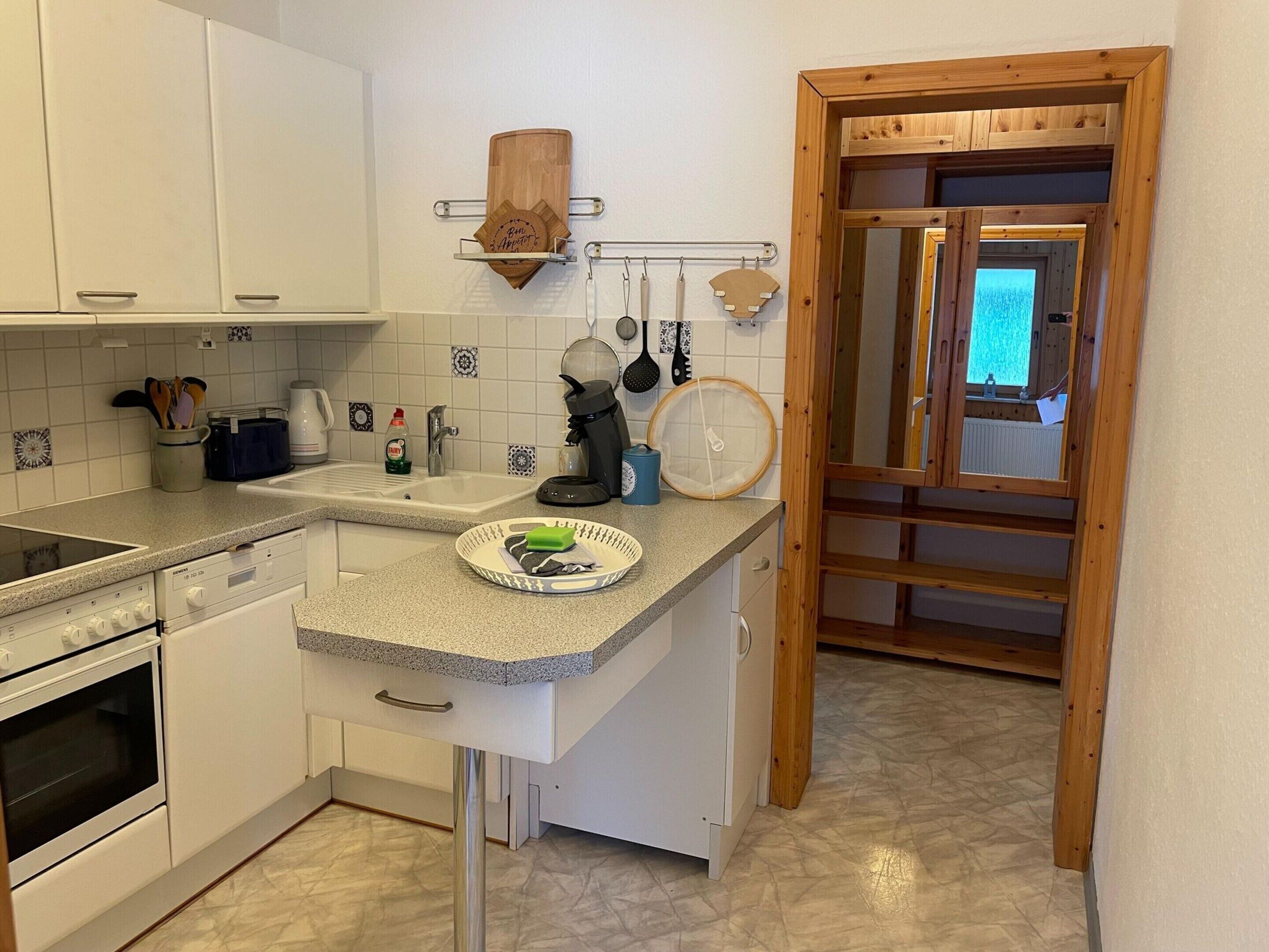 A compact kitchen with white cabinets, a granite countertop, a sink, and a stove. There is a wooden door frame and a view of a room with a window.