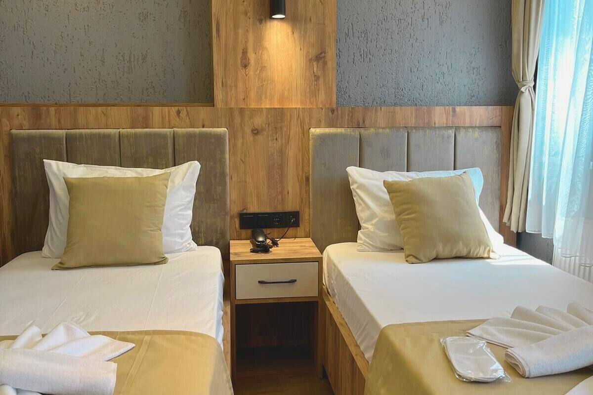 Habitación de hotel con dos camas, cabeceros de madera, una mesita de noche con lámpara y vistas al exterior.