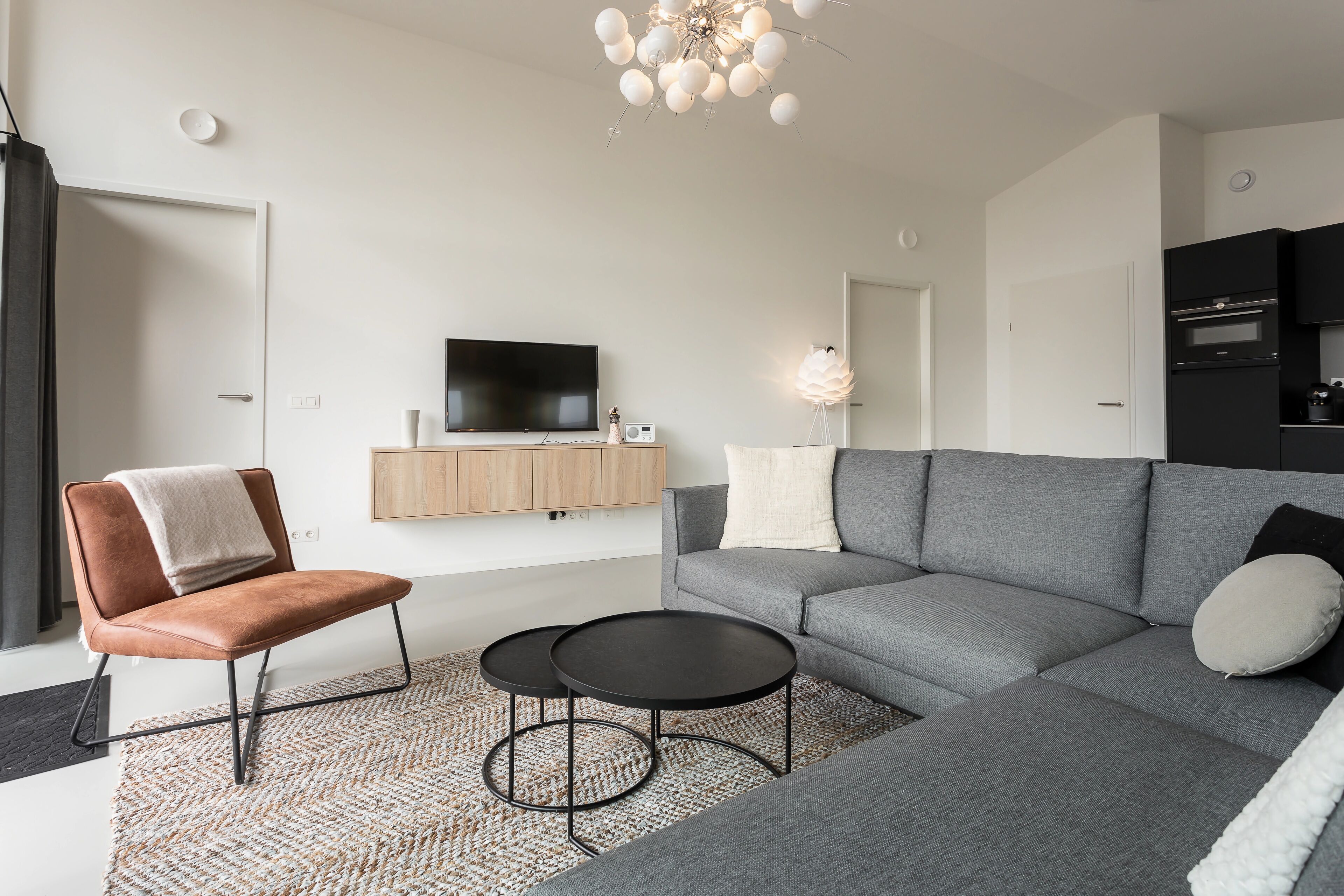 Una sala de estar moderna con un sofá gris, un sillón de cuero marrón, dos mesas de centro negras, un televisor de pantalla plana y una lámpara decorativa.