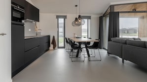 A modern kitchen with dark cabinetry, a dining area with a wooden table and black chairs, and a gray sofa in a living area with large windows. - Cottage in Wissenkerke Near Nature Reserve (Wissenkerke)
