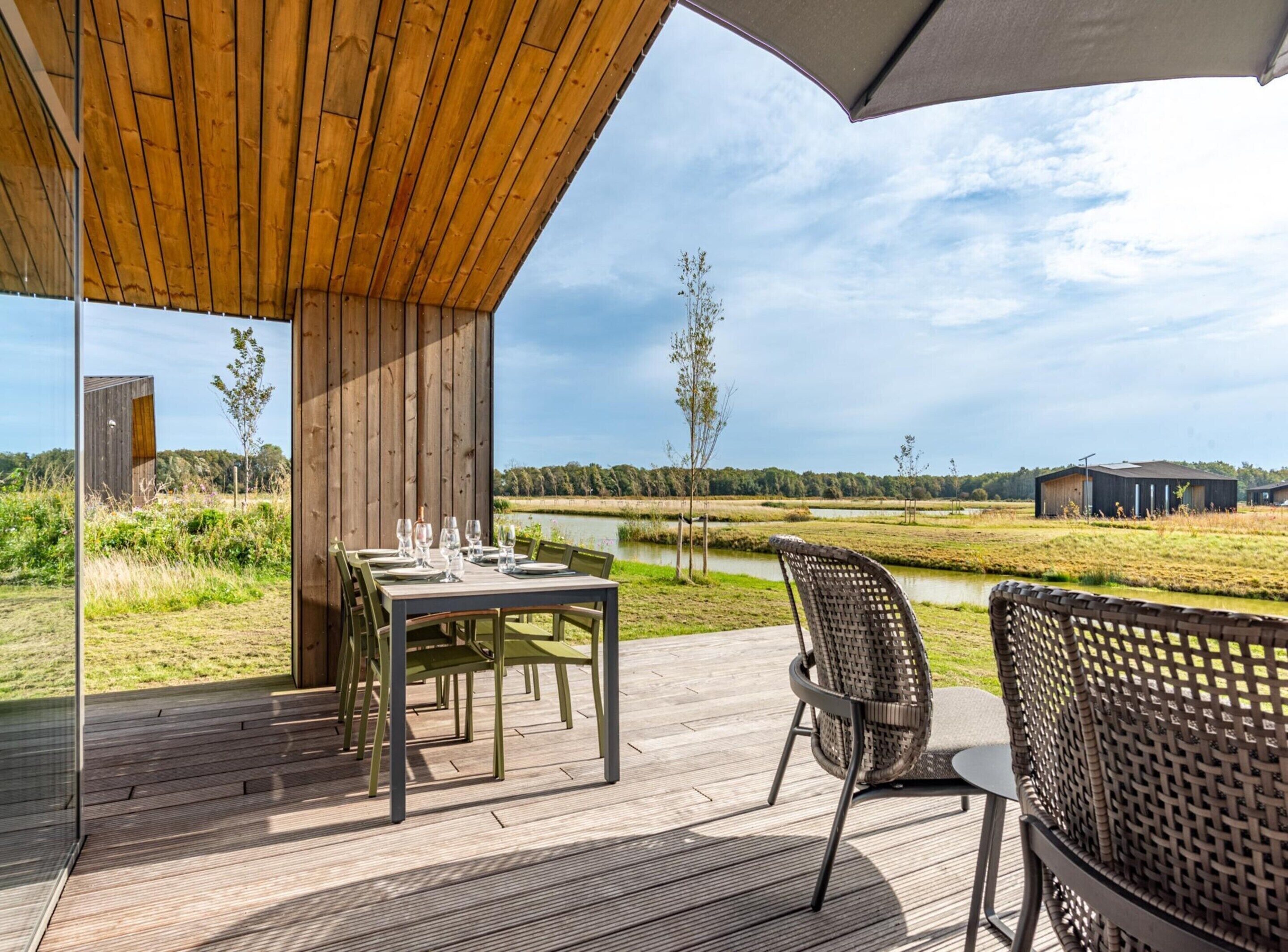 Een buitenterras met houten vloerdelen, een tafel voor vier personen en uitzicht op een meer en gebouwen in de verte.