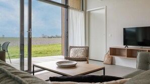 Una sala de estar moderna con un gran ventanal corredizo que da a un área exterior cubierta de césped, un televisor de pantalla plana y un sillón de mimbre.