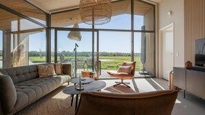 A modern living room with a sofa, armchair, and a dining table. Large windows offer a view of the outdoors. - Scenic Cottage w/ Garden (Wissenkerke)