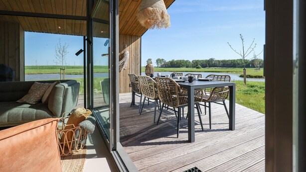 A modern outdoor dining area with a table and chairs, overlooking a body of water and open fields. - Scenic Cottage w/ Garden (Wissenkerke)