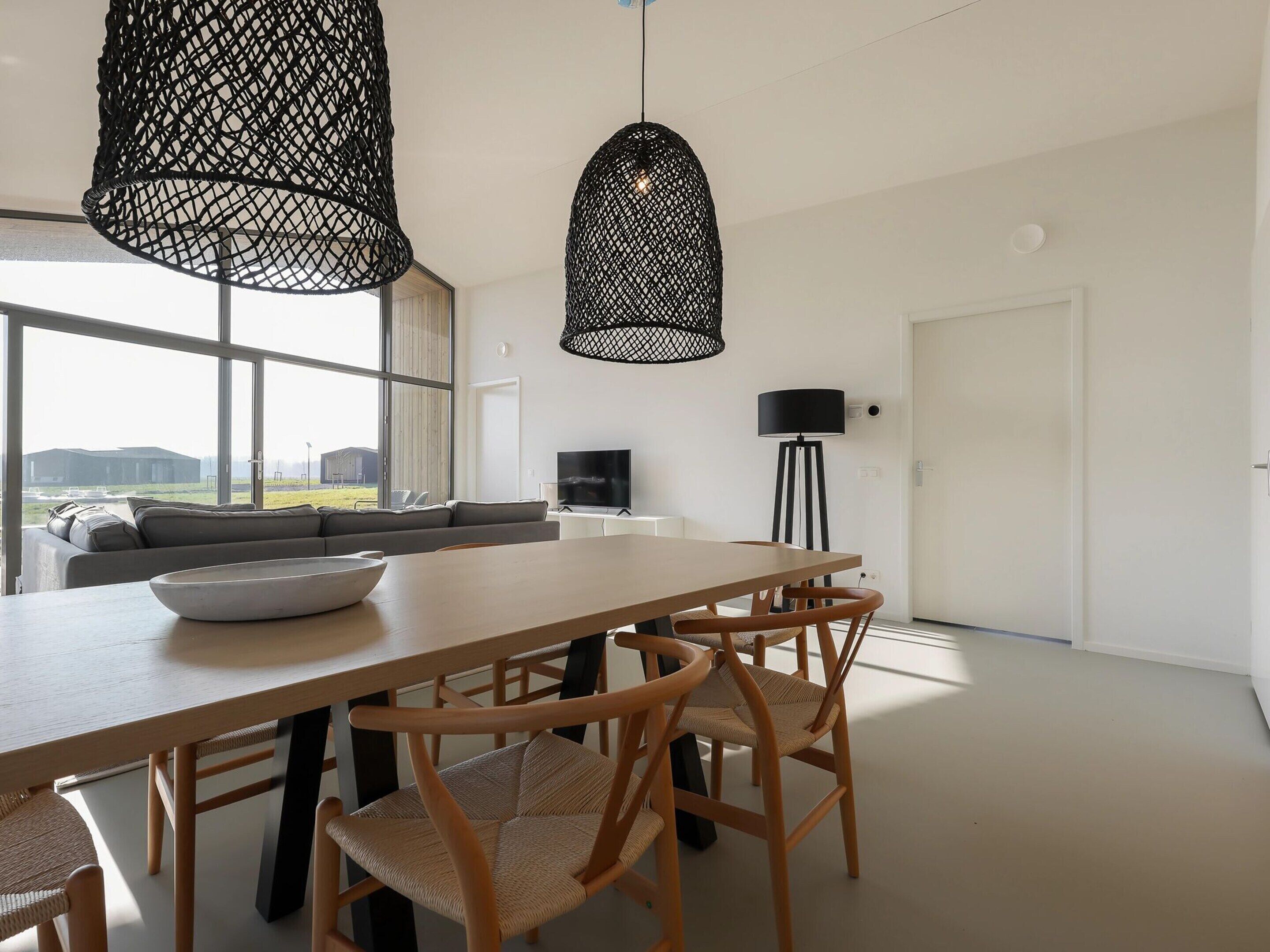 A modern dining area with a wooden table and woven chairs, a black pendant light, and a view of the outdoors through large windows.