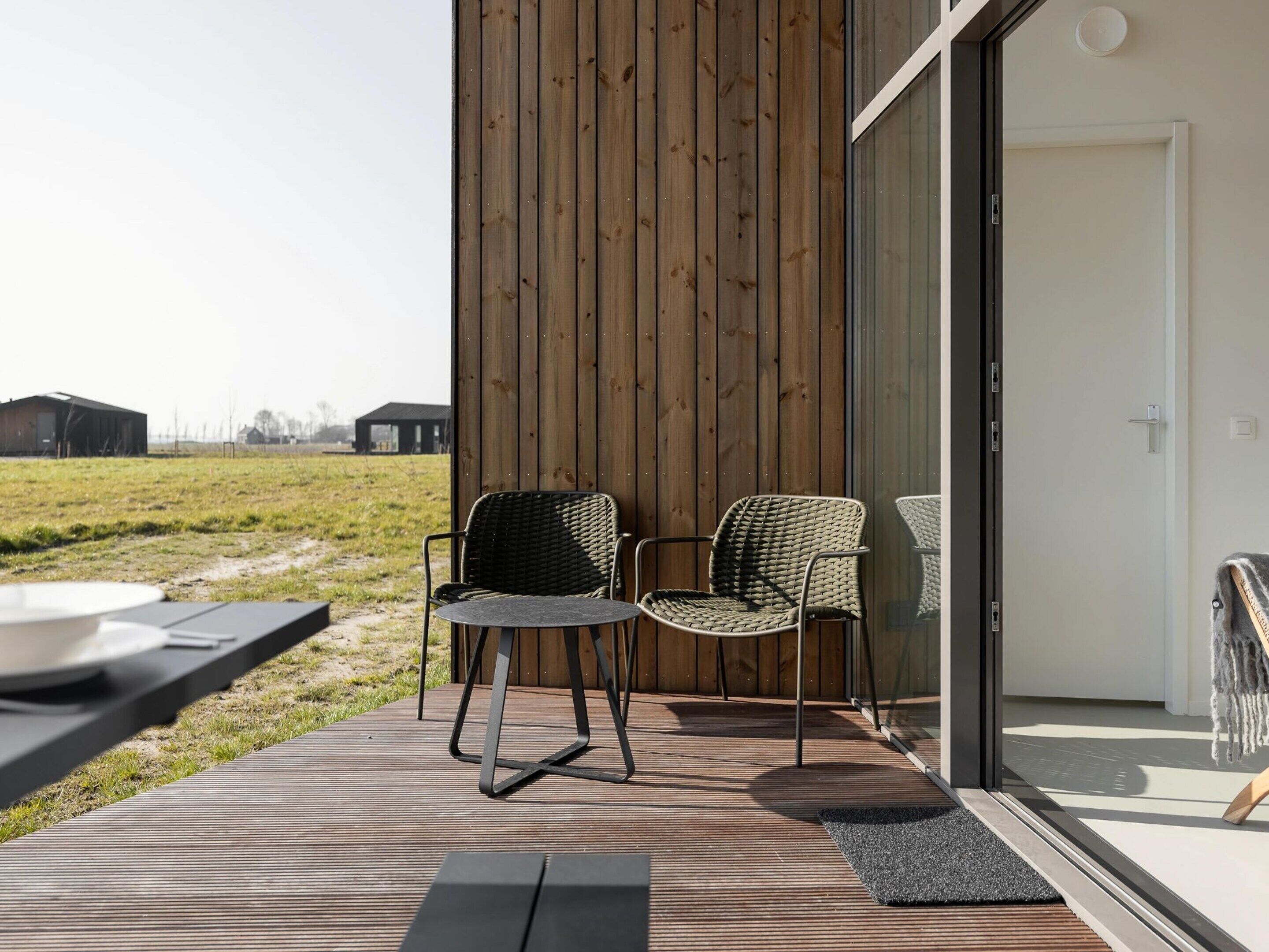 Eine Terrasse im Freien mit zwei Korbstühlen, einem kleinen Tisch, einem Holzboden und einer Glastür, die zu einem Raum führt.