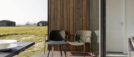 Une terrasse extérieure avec deux fauteuils en osier et une petite table, une terrasse en bois et une porte vitrée donnant sur une pièce.