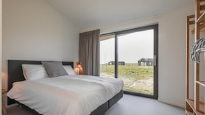 A modern bedroom with a large bed, a bedside table, a lamp, and a view of the outdoors through a sliding glass door. - Holiday Home Wissenkerke Near Nature Reserve (Wissenkerke)