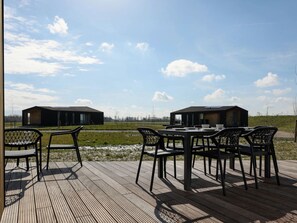 A wooden deck with a table and chairs, overlooking a grassy field and two modern buildings. - Holiday Home Wissenkerke Near Nature Reserve (Wissenkerke)