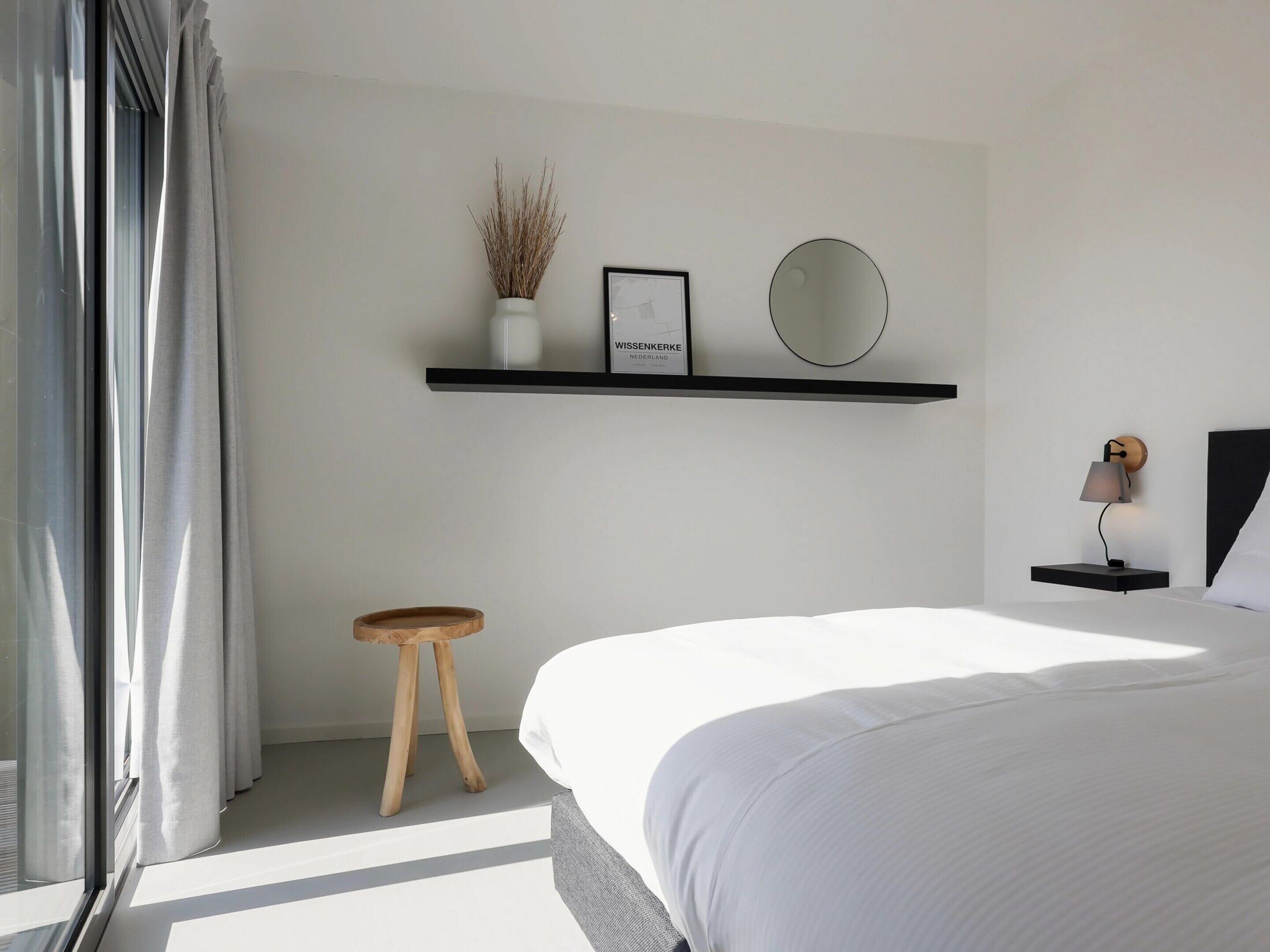 A modern bedroom with a bed, a small wooden stool, a nightstand with a lamp, a shelf with decorative items, and a window with curtains.