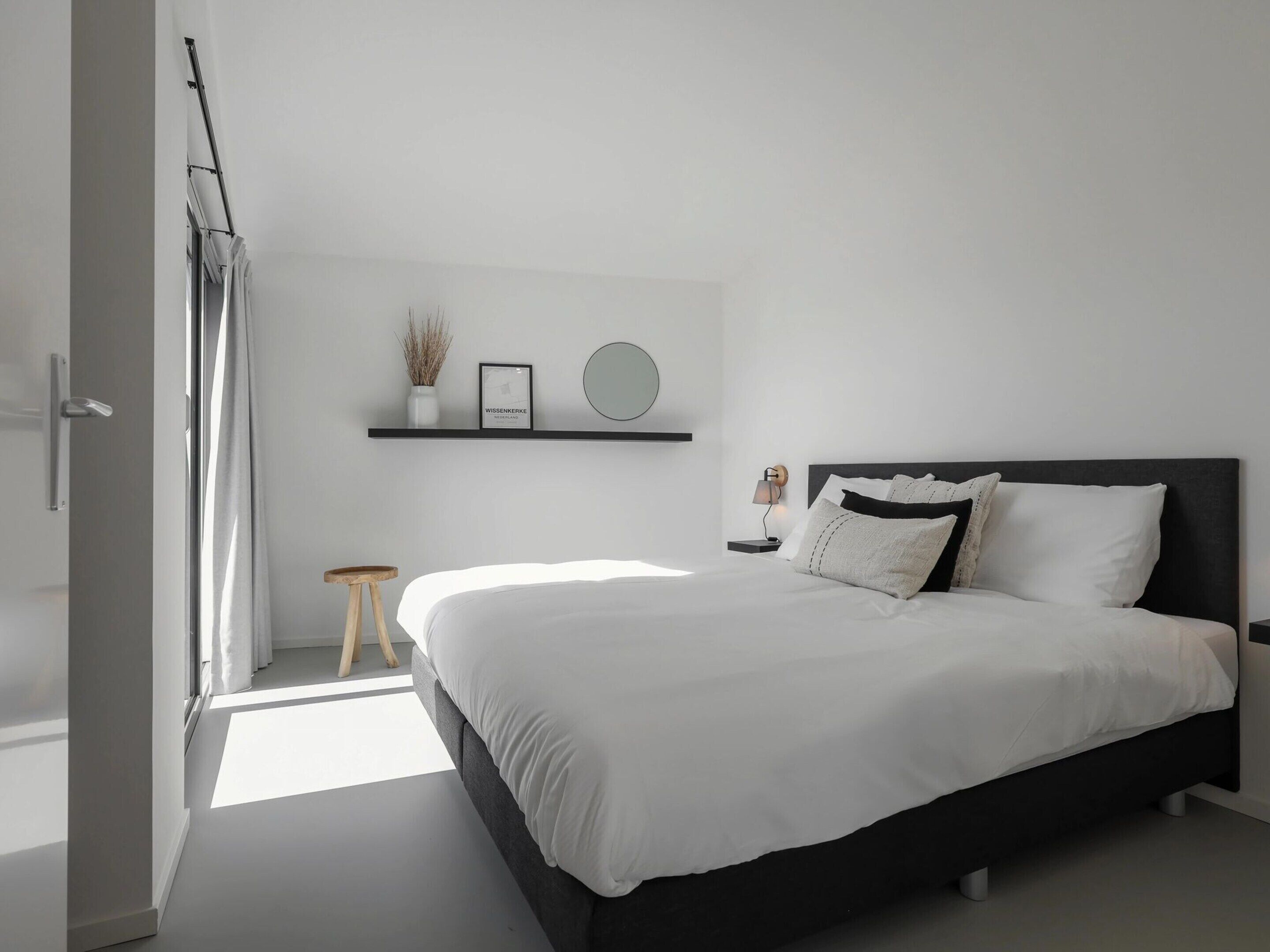 Een moderne slaapkamer met een groot bed, een nachtkastje met een lamp, een klein krukje, een plank met decoratieve spullen, en een deur met gordijnen.