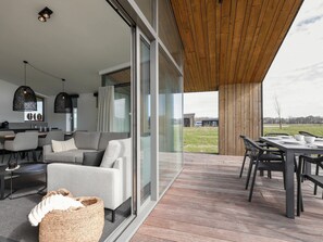 Ein moderner Wohnbereich mit Sofa, Esstisch und Stühlen. Es gibt eine Küchenzeile mit Arbeitsfläche und Schränken. Der Raum hat große Fenster und Blick ins Freie.