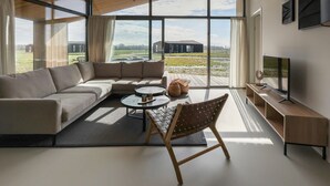 A modern living room with a large sectional sofa, a round coffee table, and a wooden chair. There is a television on a wooden stand and a large window offering a view of the outdoors. - Holiday Home Wissenkerke Near Nature Reserve (Wissenkerke)