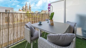 Un balcón con mobiliario de mimbre, una mesa con una taza y una planta, y una vista de un castillo al fondo.