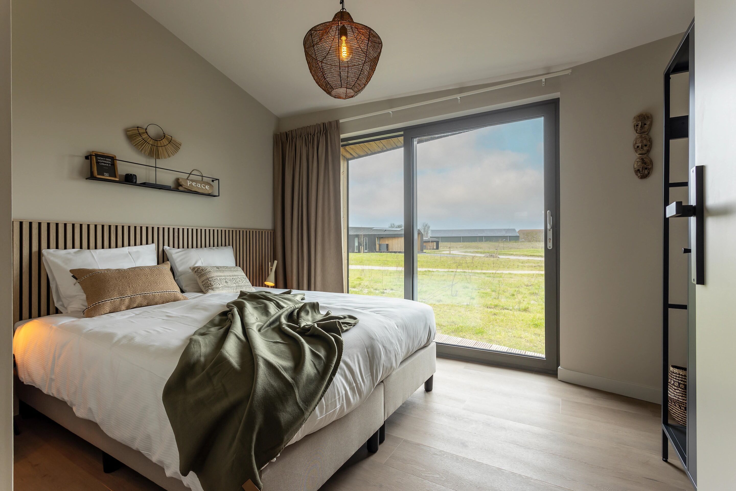 A bedroom with a large bed, a hanging light fixture, a view through a glass door, and a shelf with decorative items.