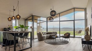 Un amplio espacio habitable con un gran ventanal que ofrece vistas a un lago, un comedor con una mesa y sillas modernas, y una zona de estar con un sofá y una mesa de centro.