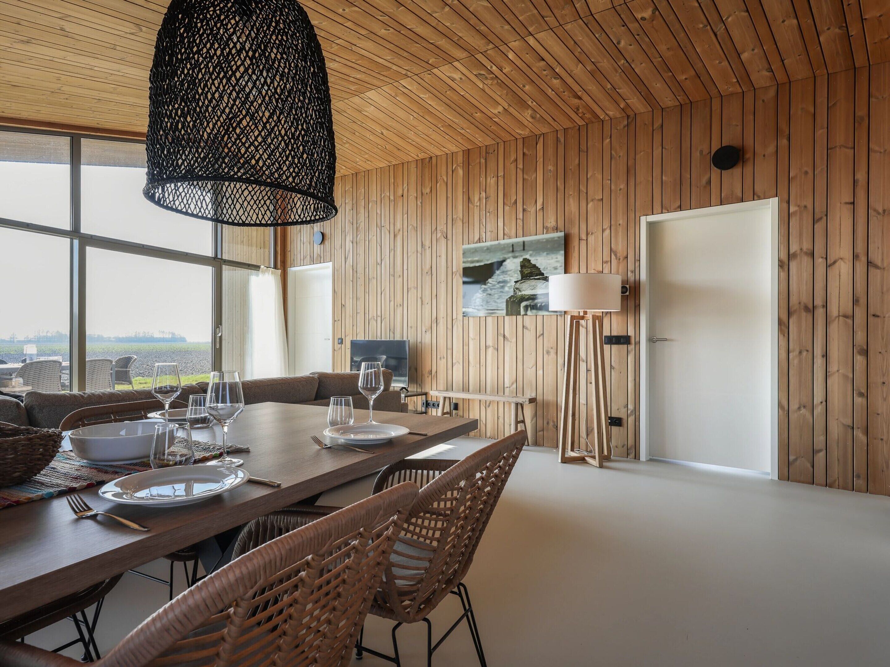 Un'area pranzo con un tavolo di legno apparecchiato per quattro persone, circondato da sedie in vimini. Una grande finestra offre vista sull'esterno.