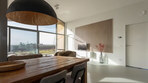 A modern dining area with a wooden table and chairs, a large pendant light, and a view of the countryside through large windows. - Cottage in Wissenkerke Near Het Bokkegat (Wissenkerke)