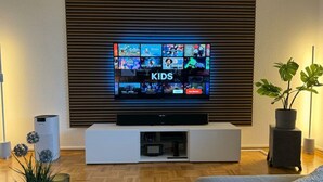 A living room with a flat-screen TV mounted on a wall, a white entertainment unit below, and a potted plant on the right. - Lovely Apartment Near the Water (Weilburg)