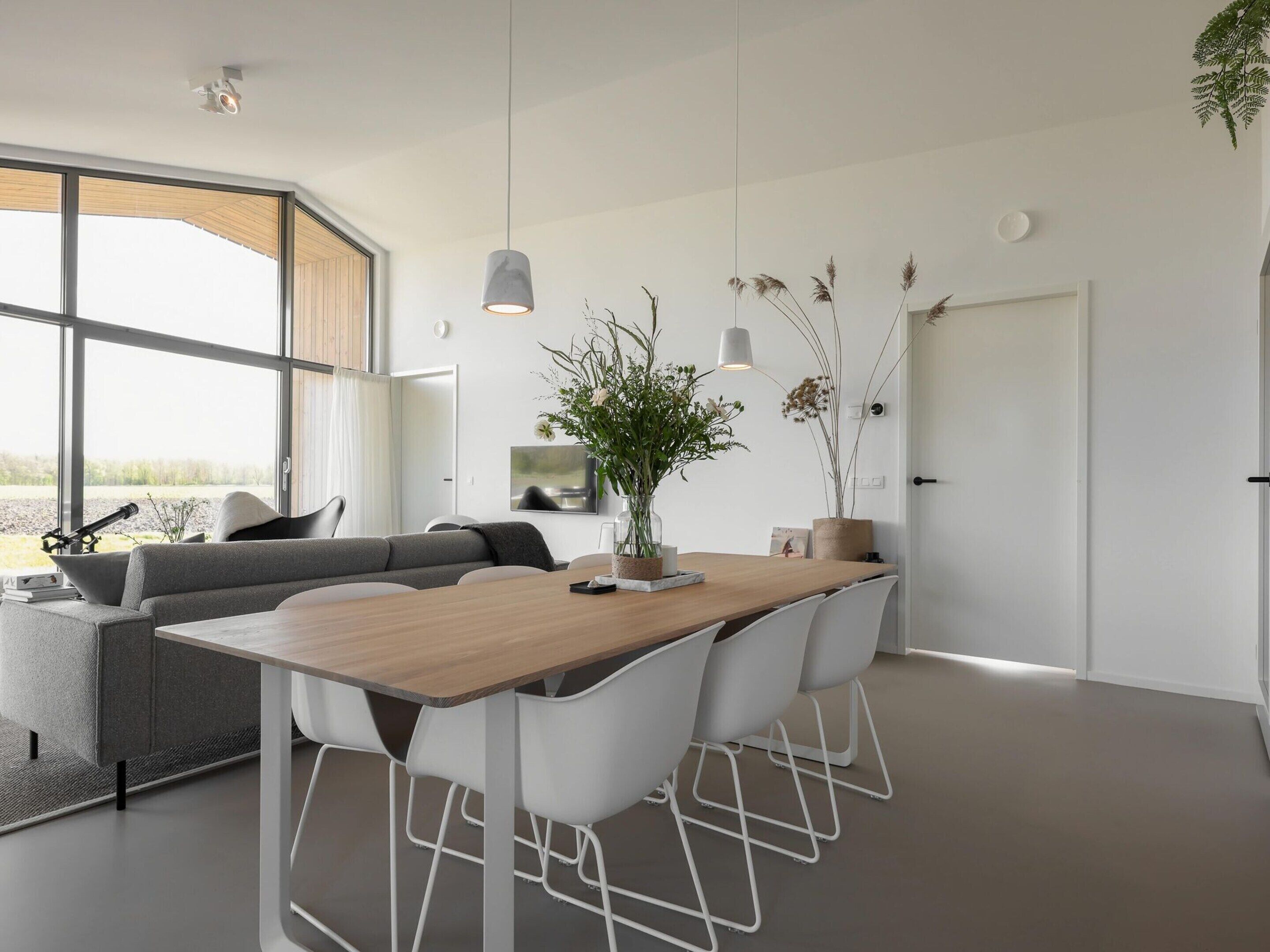 A modern living space with a wooden dining table, white chairs, a grey sofa, and a large window offering a view of the outdoors.