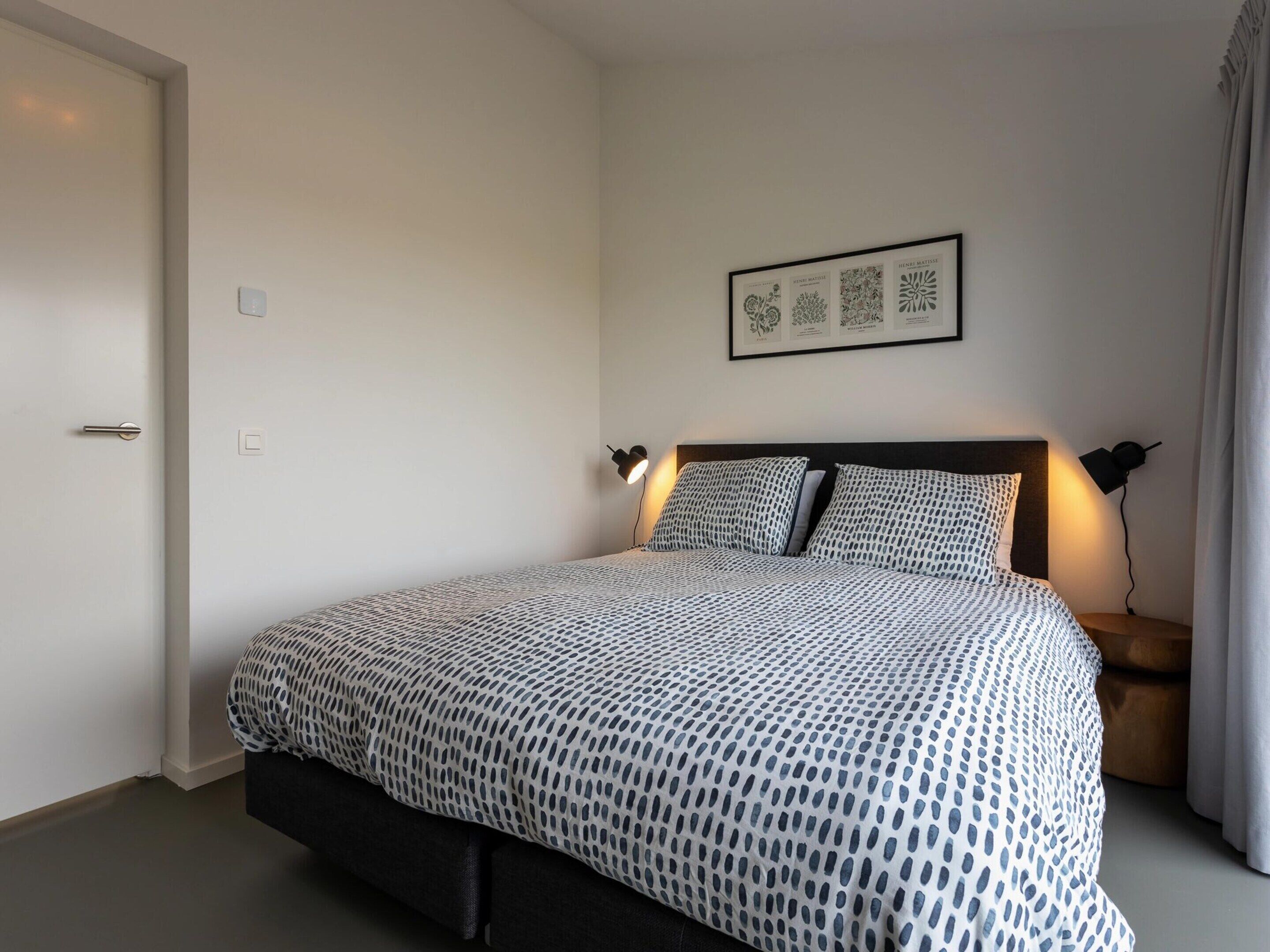 A neatly arranged bedroom with a bed, two bedside lamps, a framed botanical print on the wall, and a door.
