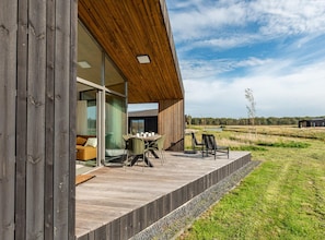 A modern building with a wooden deck, a dining area with chairs and a table, and a view of the surrounding landscape. - Modern Eco Home Near Beach (Wissenkerke)