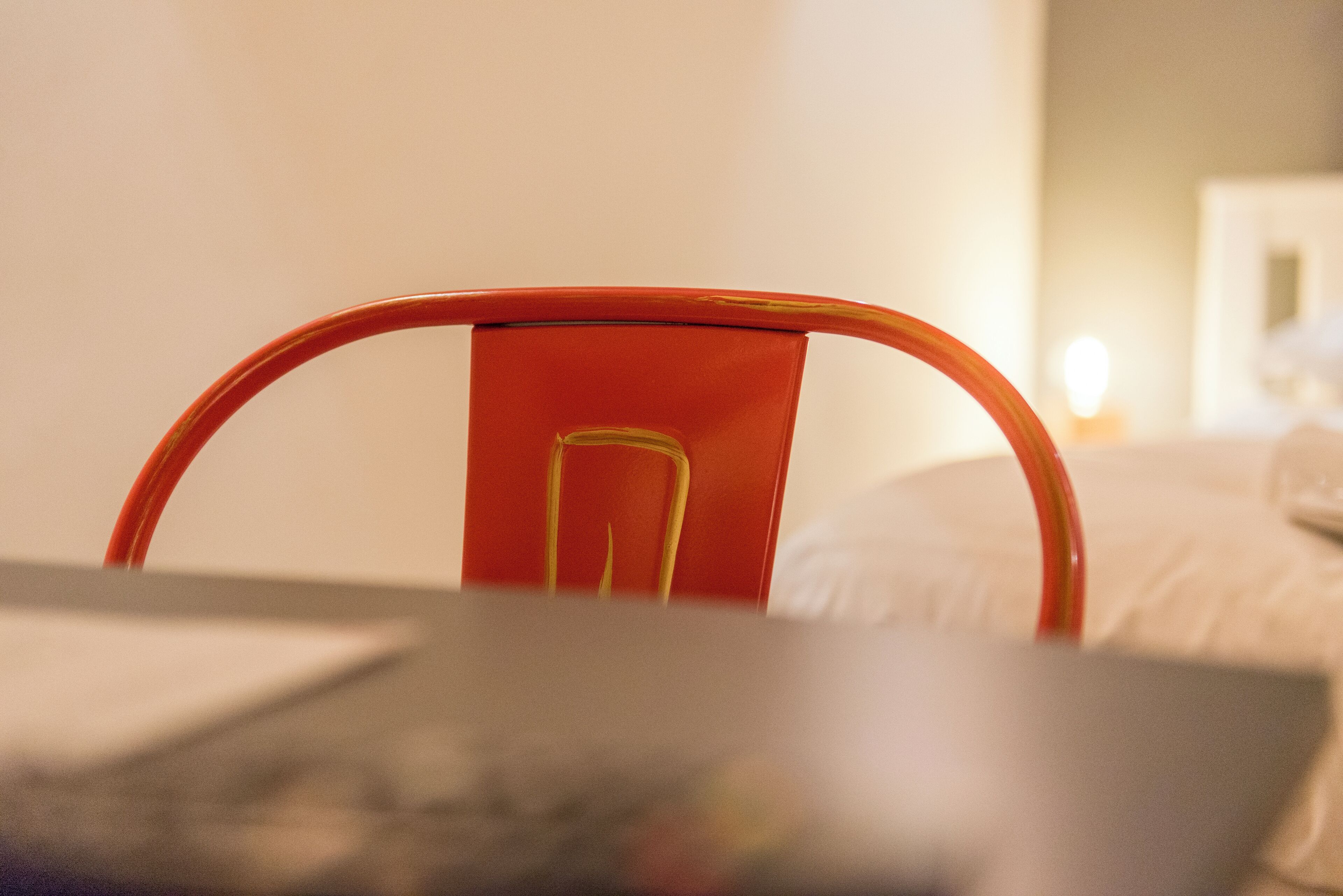 Un gros plan d’une chaise en métal rouge, dotée d’un dossier incurvé et d’un motif ajouré au centre, placée devant un lit recouvert de draps blancs.