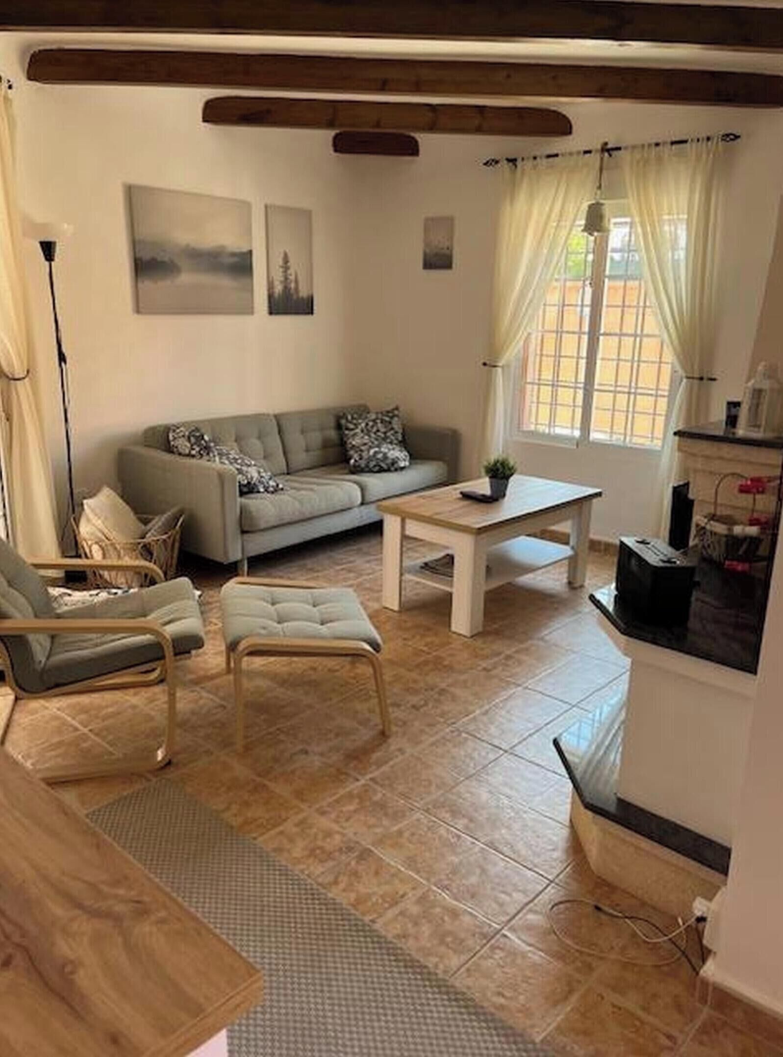 A living room with a sofa, armchair, coffee table, and a small table with a plant. There is a window with curtains, a lamp, and a wall-mounted picture.