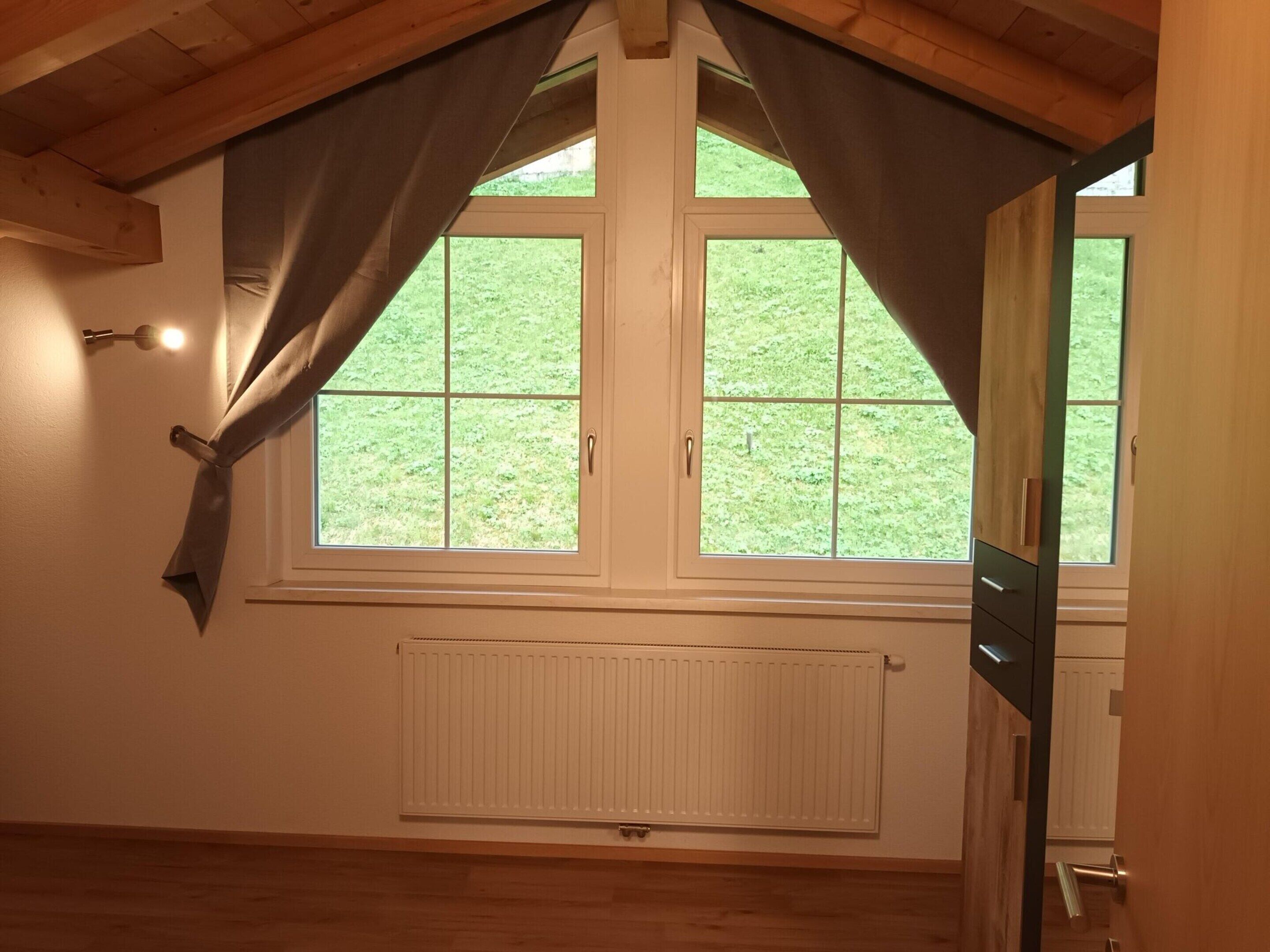 A room with a sloped ceiling, two windows with white frames, a radiator below the windows, and a wooden floor.
