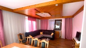 Uma sala de estar aconchegante com teto de madeira, um sofá, uma mesa de jantar com cadeiras e uma televisão sobre um móvel.
