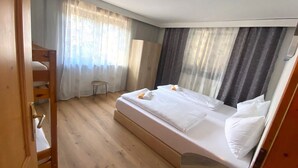 Quarto de hotel com uma cama de solteiro em madeira, um banquinho pequeno e uma janela grande com cortinas leves.